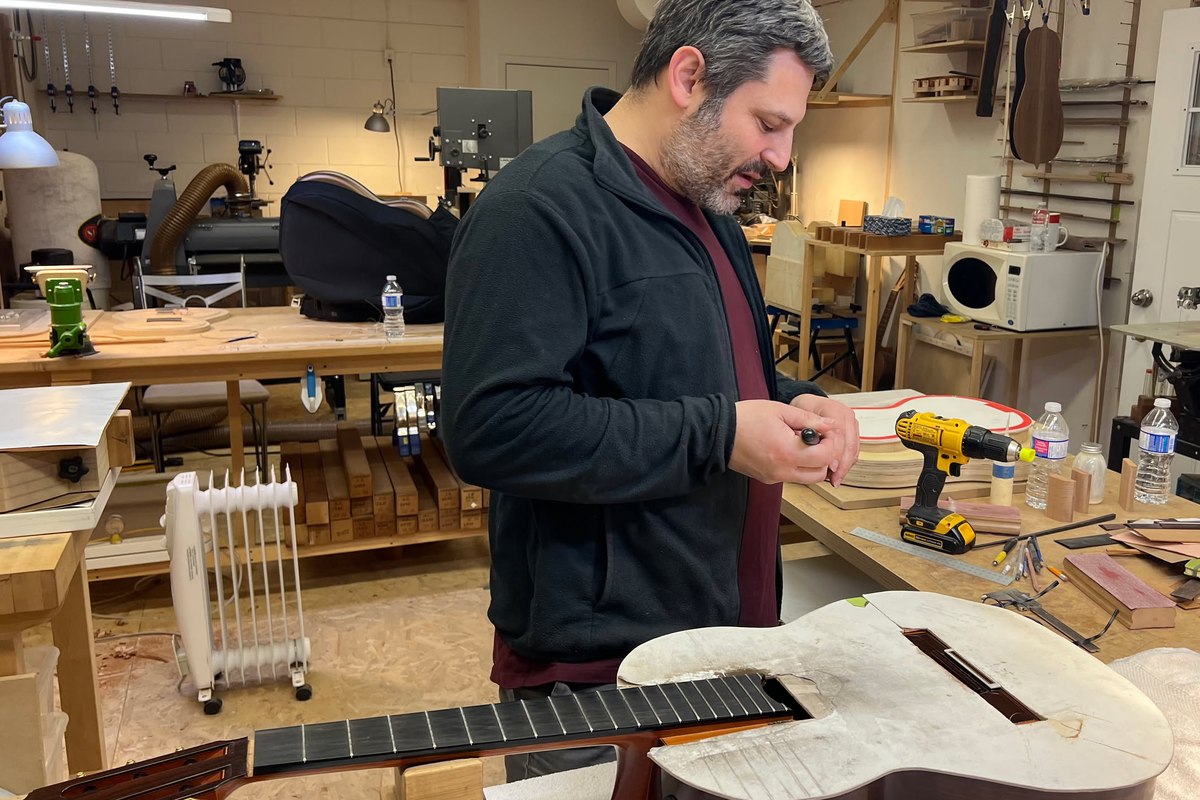 Mark Usherovich at his workbench, head bent over his hands, an unfinished guitar in the white lying on the bench in front of him